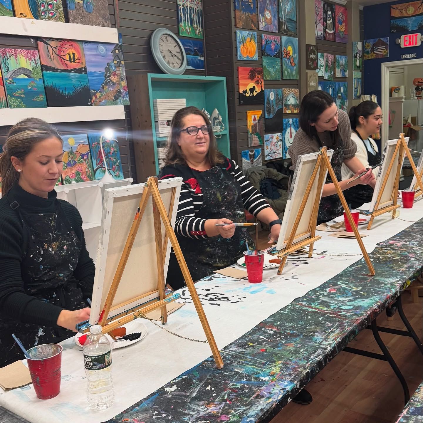 A group of women are sitting at a long table painting on canvases.