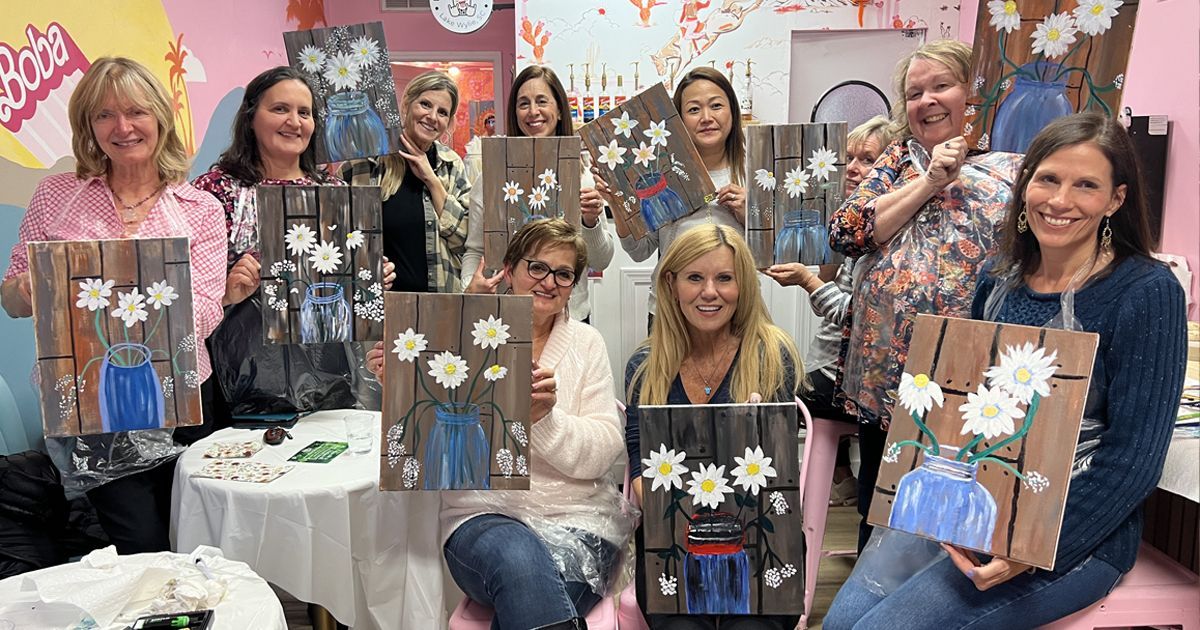 Group of people holding framed flower art pieces in a craft workshop, smiling in a bright room
