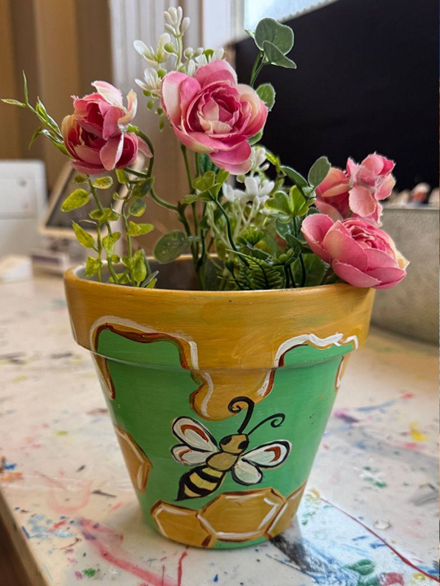 Painted flower pot with bee and honeycomb design, topped with pink roses and greenery.