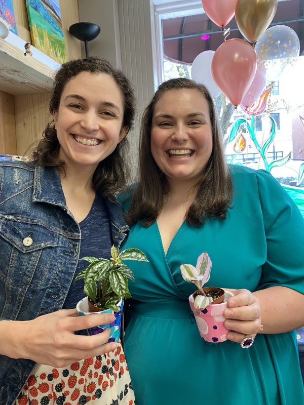 Two smiling women holding small potted plants at a party with pink balloons and decorations