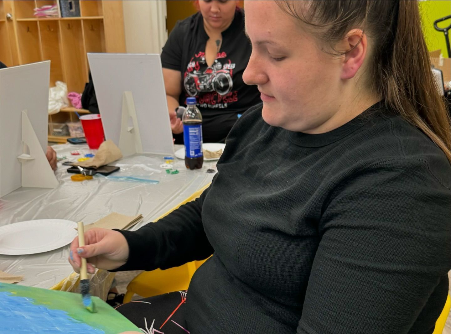 A woman is sitting at a table painting a picture with a brush.