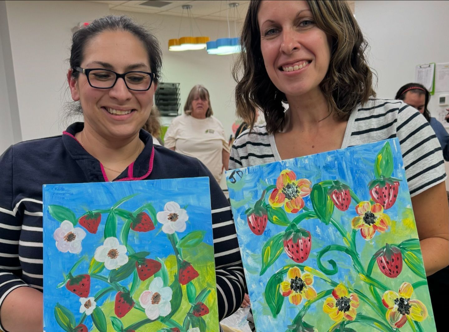 Two women are holding up paintings of strawberries and flowers.