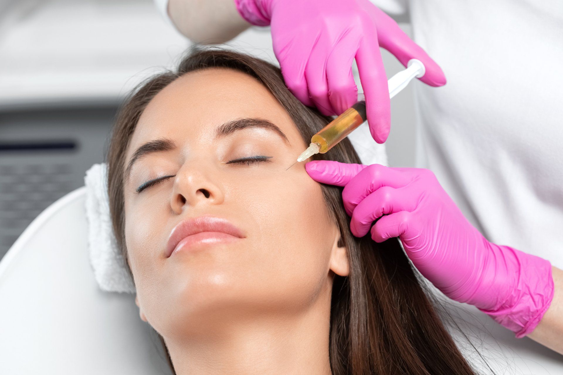 Aesthetic treatment with pink-gloved hands applying a syringe to a woman's forehead.