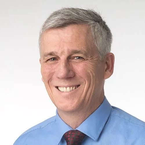 Smiling man in a blue shirt and red tie, against a light gray background.