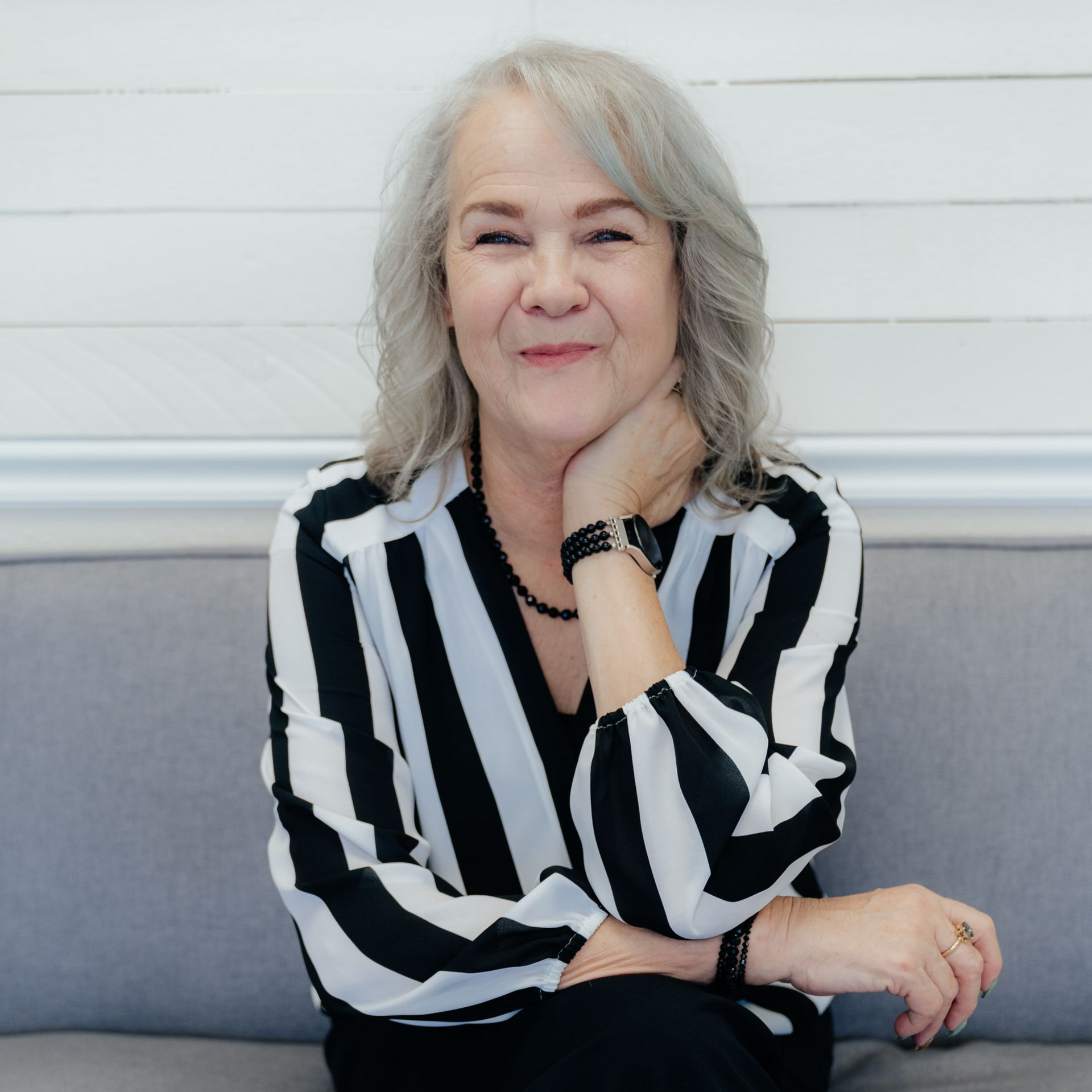 Woman with graying blonde hair, smiling, wearing a black patterned shirt, against a plain wall.