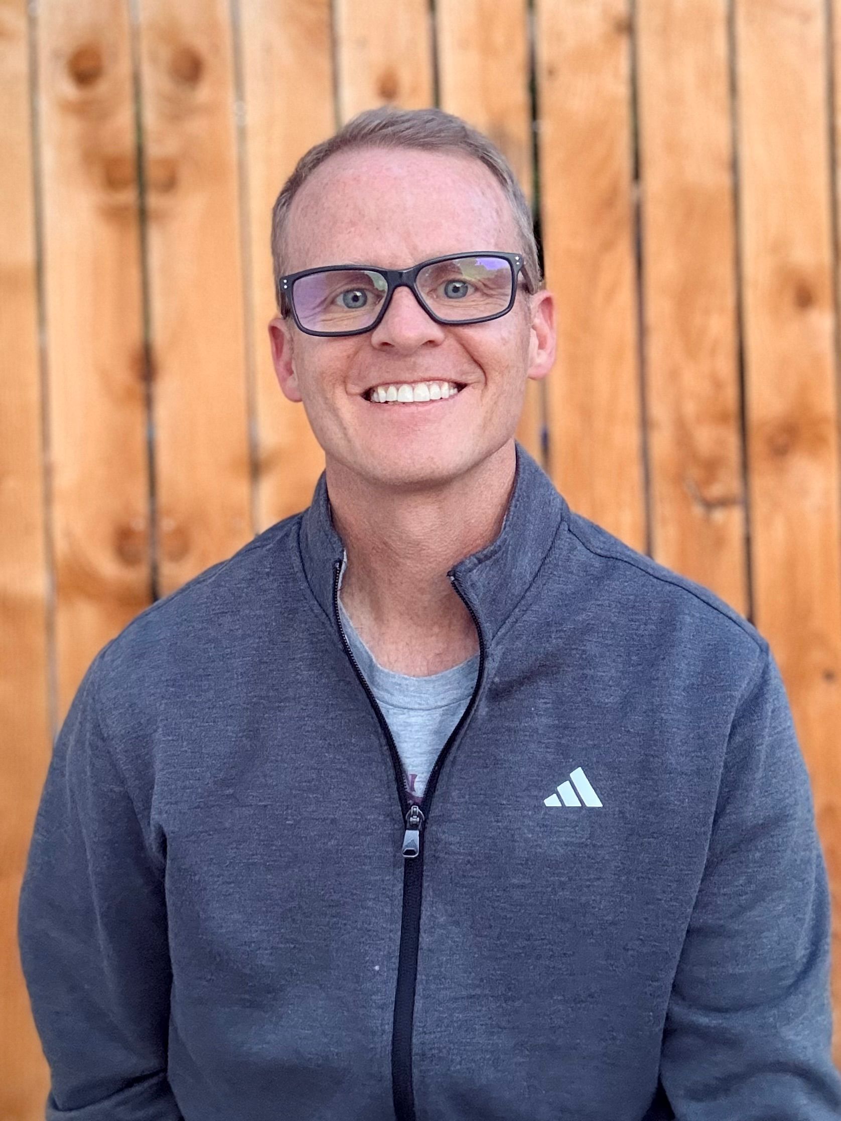 Smiling person in glasses wearing a blue jacket, standing in front of a wooden wall.