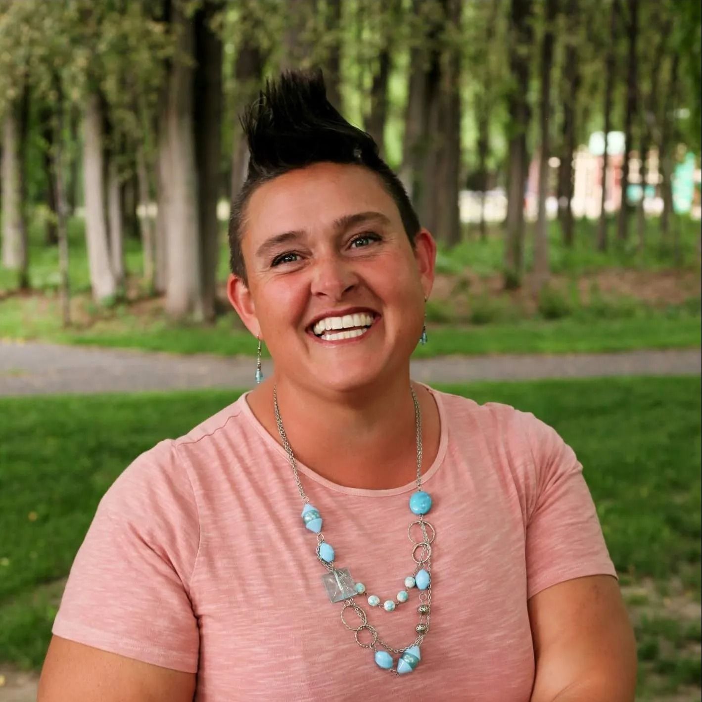 Woman with a mohawk and necklace smiles broadly in a park.