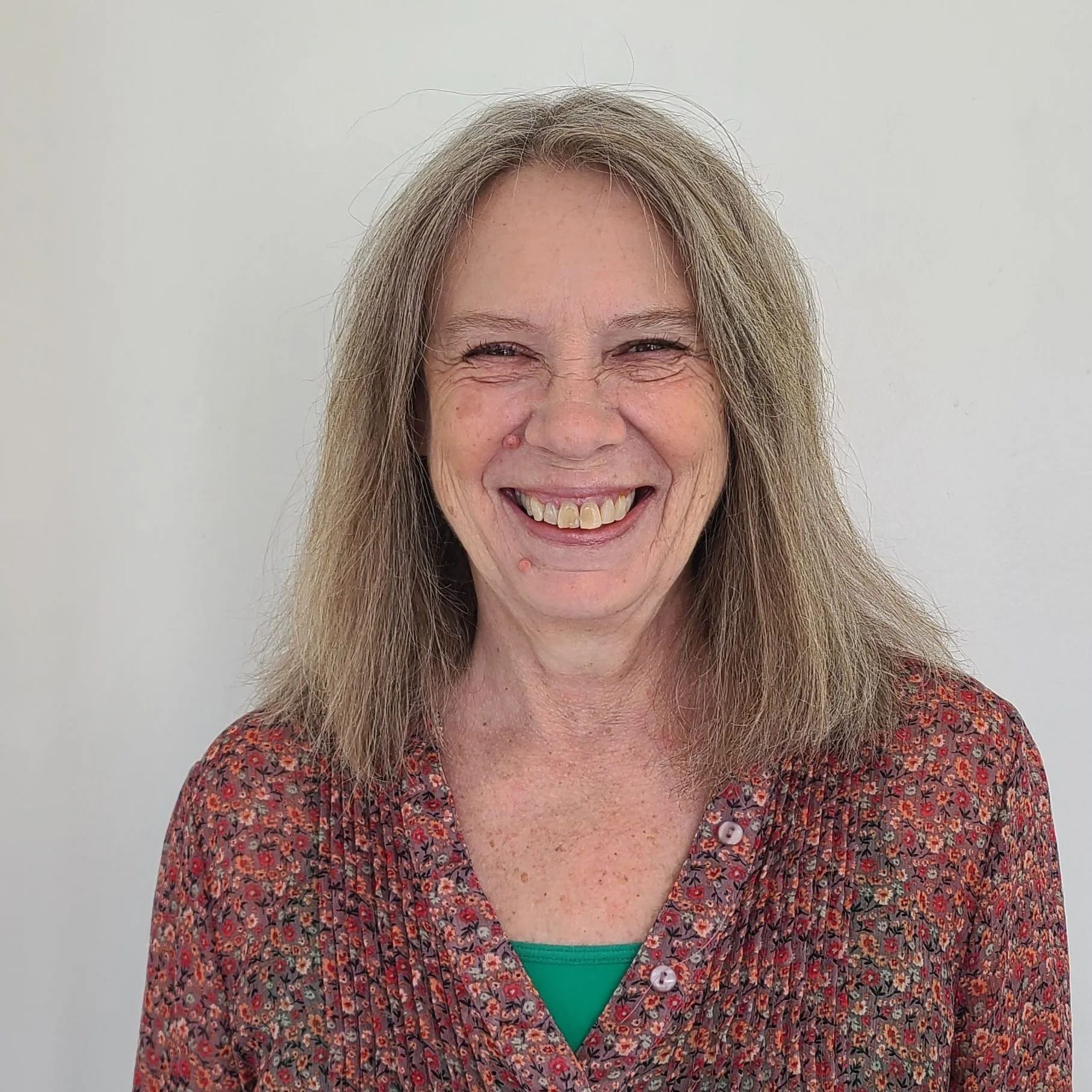 Woman with long gray hair smiles, wearing a floral shirt and green undershirt, against a white wall.
