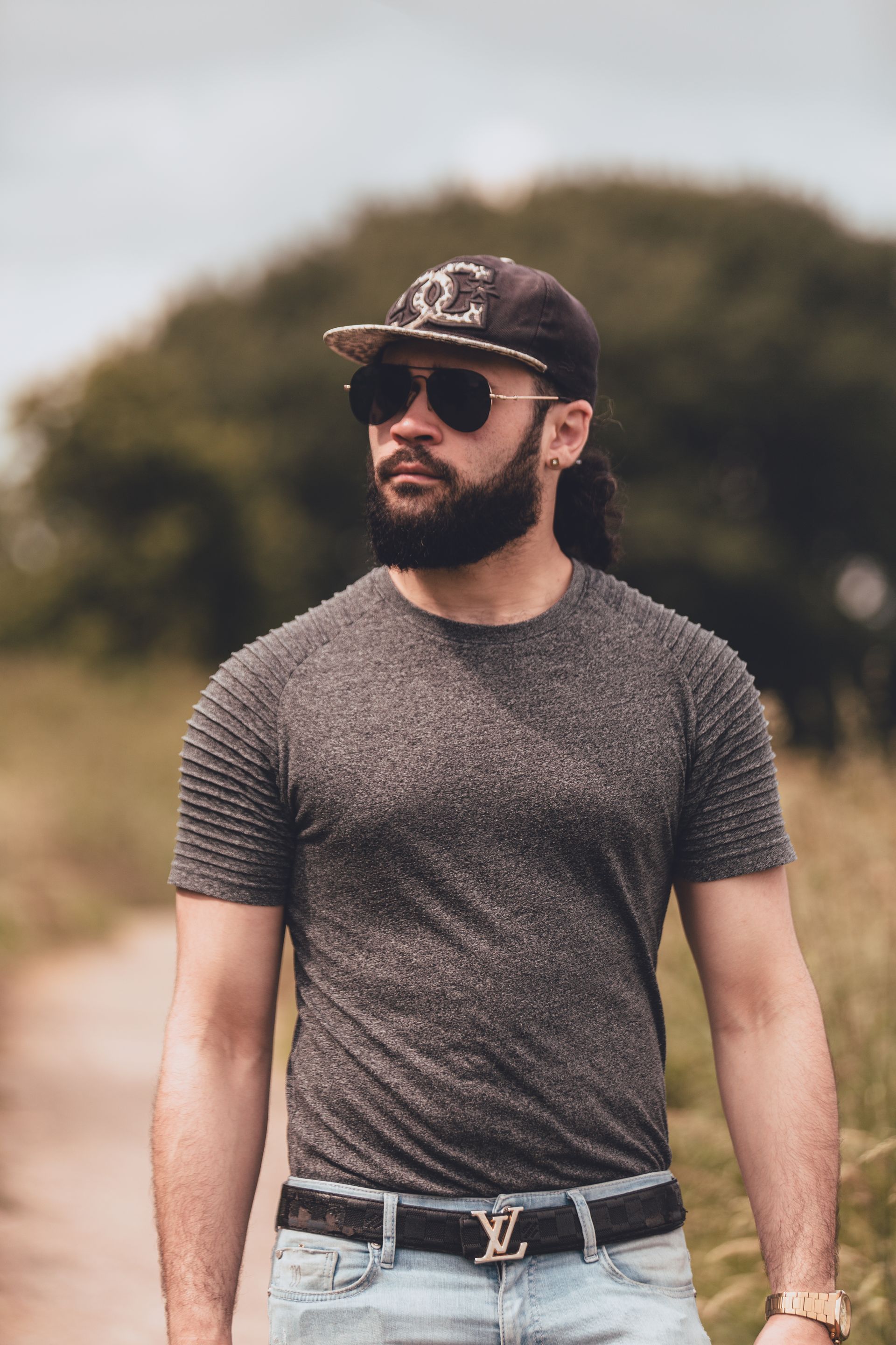 Een man loopt buiten op een pad, omringd door natuur. Hij draagt een donkere pet met een logo, een donkere zonnebril, een donker t-shirt met geribbelde mouwen, een zwarte riem met een zilveren gesp en lichtblauwe jeans. De man heeft een volle baard en een oorbel in zijn linkeroor. In de achtergrond zijn bomen en een bewolkte hemel zichtbaar.