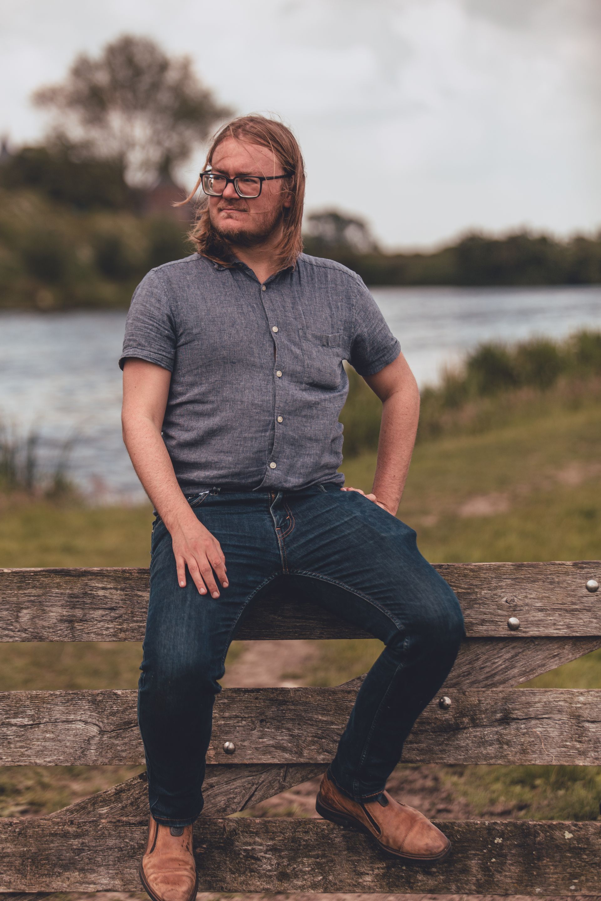 Een man zit op een houten hek bij een waterlichaam, met groene vegetatie op de achtergrond. Hij draagt een bril, een blauw overhemd met korte mouwen, een spijkerbroek en bruine schoenen. De man kijkt in de verte en heeft zijn handen op zijn heupen geplaatst. Achter hem is een boom en wat lijkt op een kasteel of groot gebouw in de verte zichtbaar.