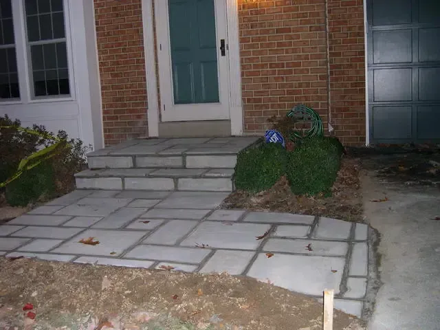 Front steps and walkway to a brick house, with uneven concrete slabs and a green front door.
