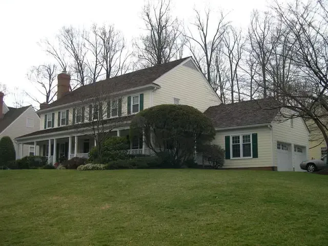 Large beige two-story house with white trim and a front porch on a grassy lawn, surrounded by bare trees