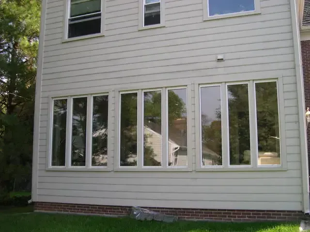 Side view of a beige house with large white-framed windows and a brick foundation