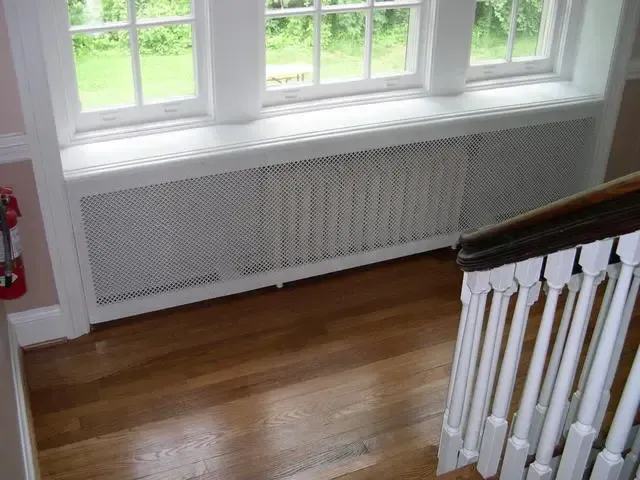 White radiator cover beneath windows in a bright room with hardwood floor and stair railing