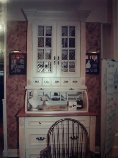 White hutch desk with drawers and chair in a patterned room, framed by floral wallpaper and wall art.