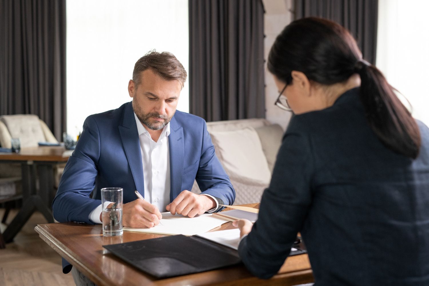 Man in blue suit signing document at a table, facing a woman in dark suit.