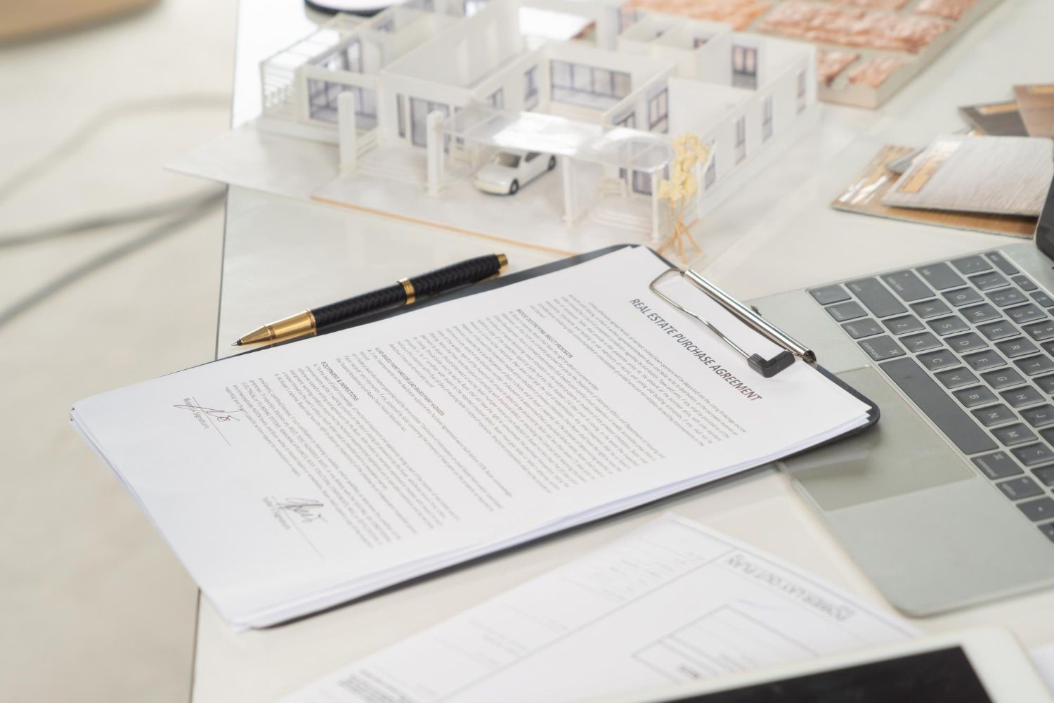 Clipboard with contract, pen, architectural model, and laptop on a desk.