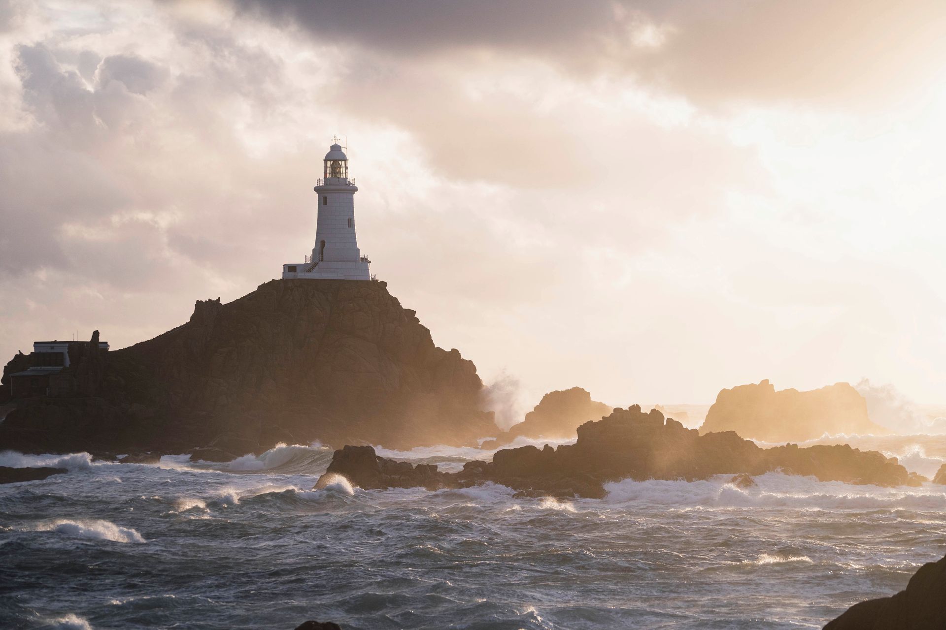 Lighthouse on rocky coast at sunset with rough waves and golden light