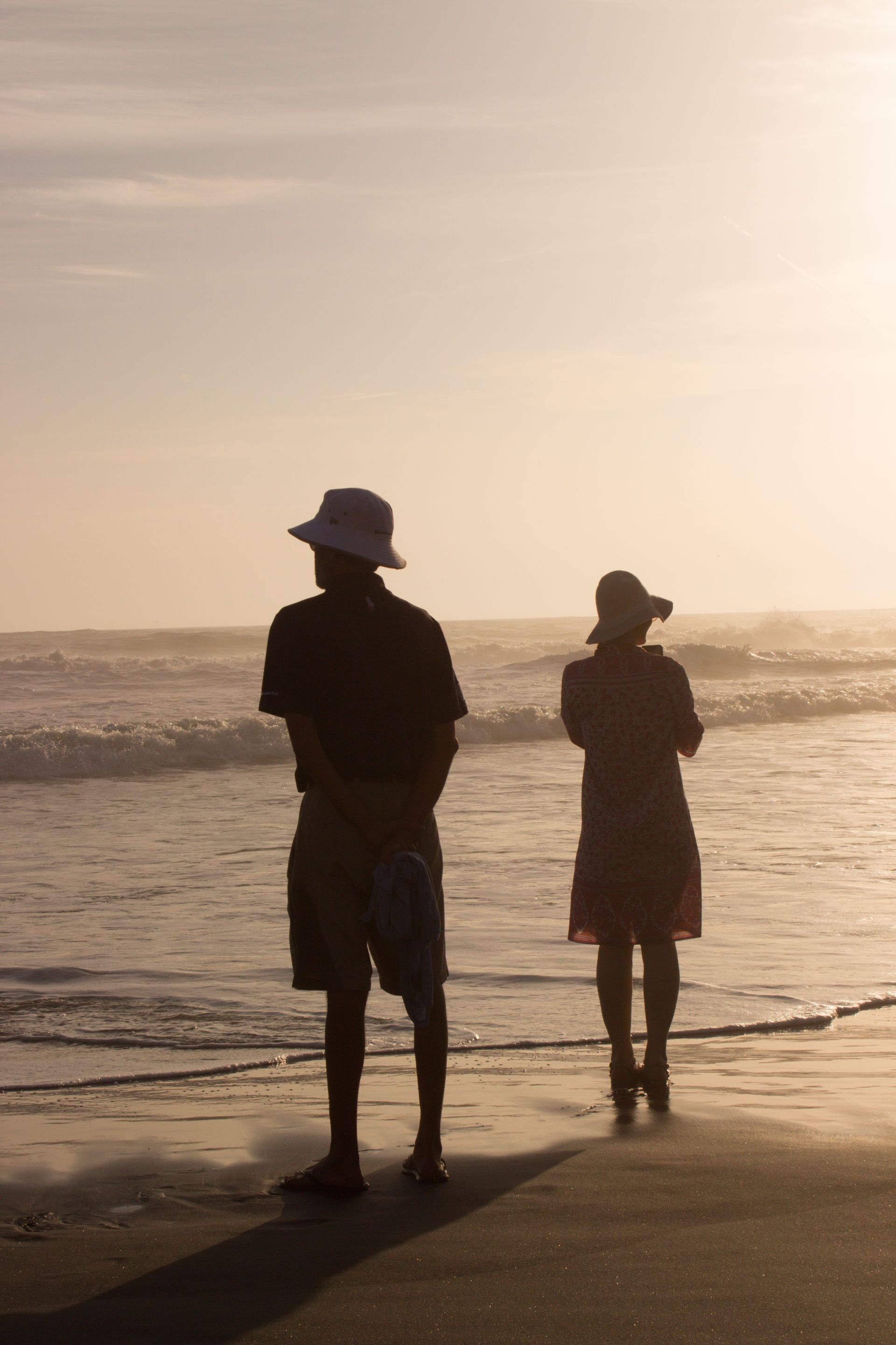 Two people stand on a beach at sunset, facing the ocean with gentle waves and warm light.