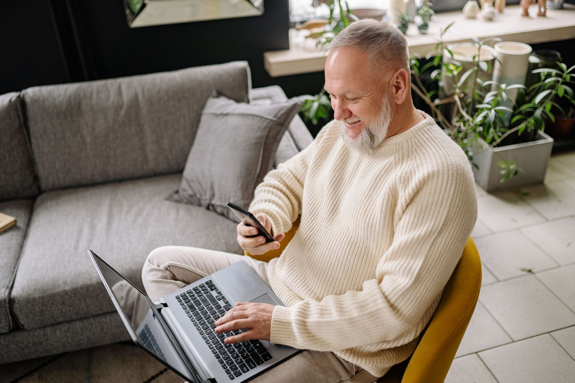 Man in cream sweater using a laptop and smartphone while seated on a yellow chair in a living room