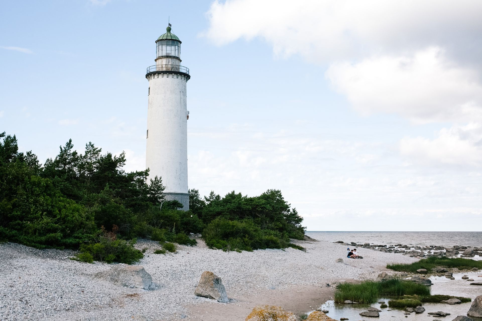 White lighthouse on a rocky shoreline with green shrubs and calm water under a cloudy sky