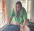 Person in a green shirt leans over a massage table in a sunlit room, hands working on the client’s back