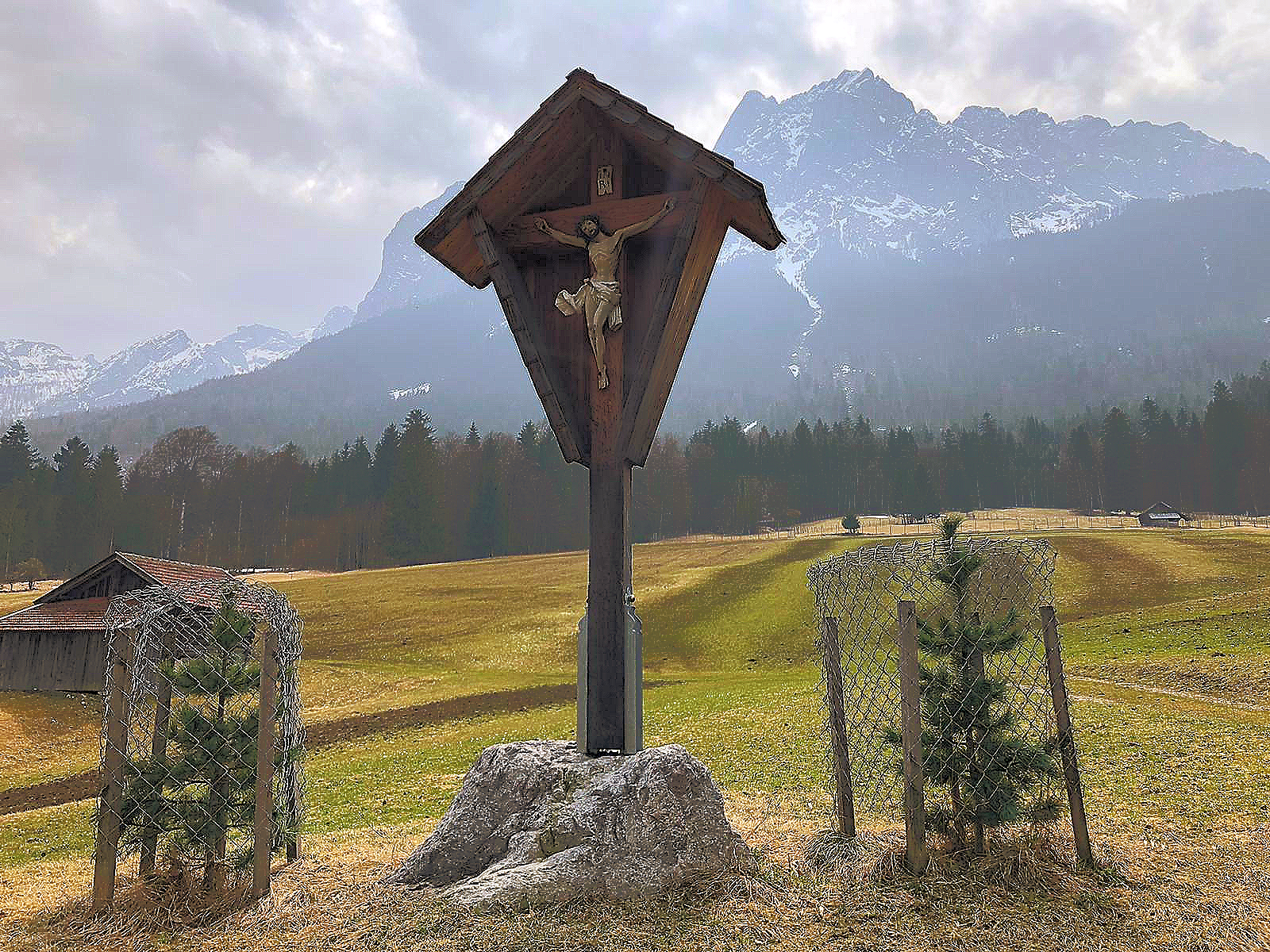 Ein Holzkreuz mit einer Jesusstatue darauf auf einem Feld mit Bergen im Hintergrund.