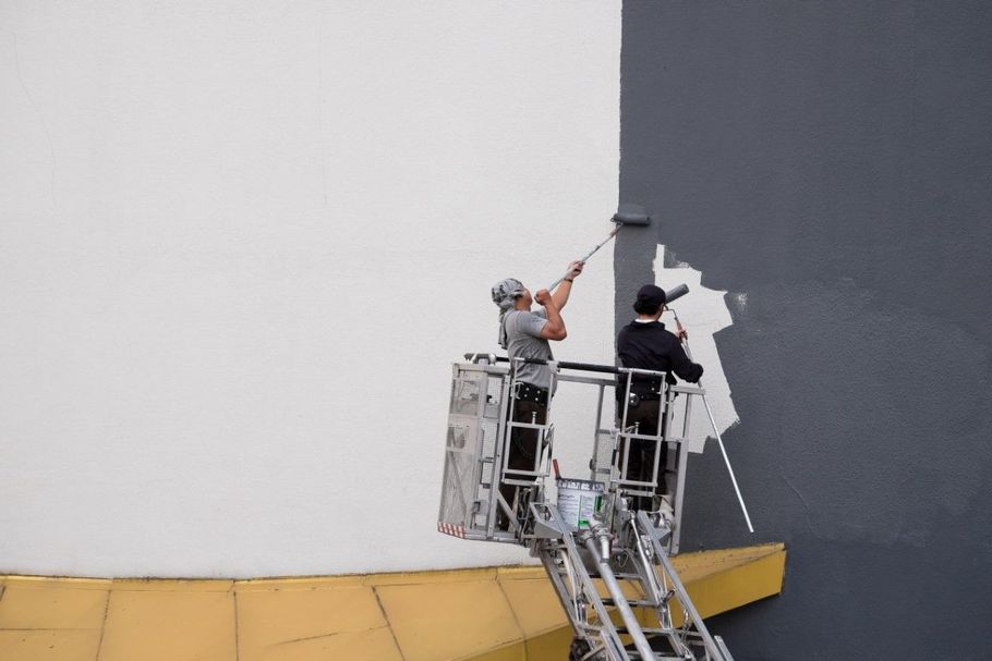 Two workers in lifts painting a tall wall, one white and one dark gray, with a large yellow base below