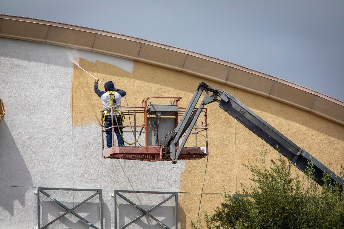 Worker painting a building exterior from a cherry picker beside a boom lift