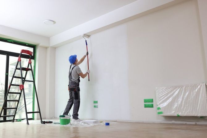 Worker painting a white wall indoors beside a ladder and green paint bucket