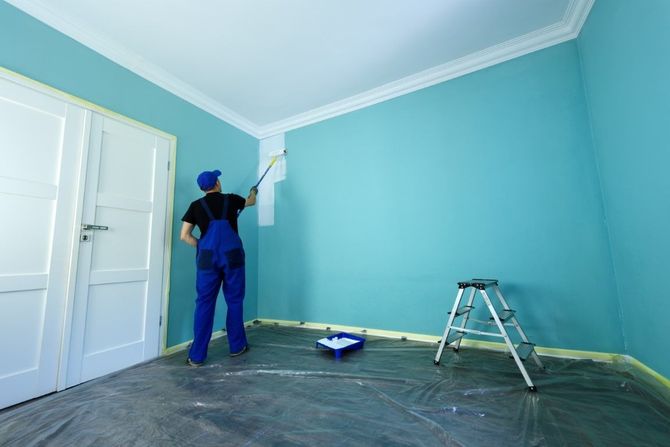 Painter painting a light blue wall in an empty room with a ladder and drop cloths.