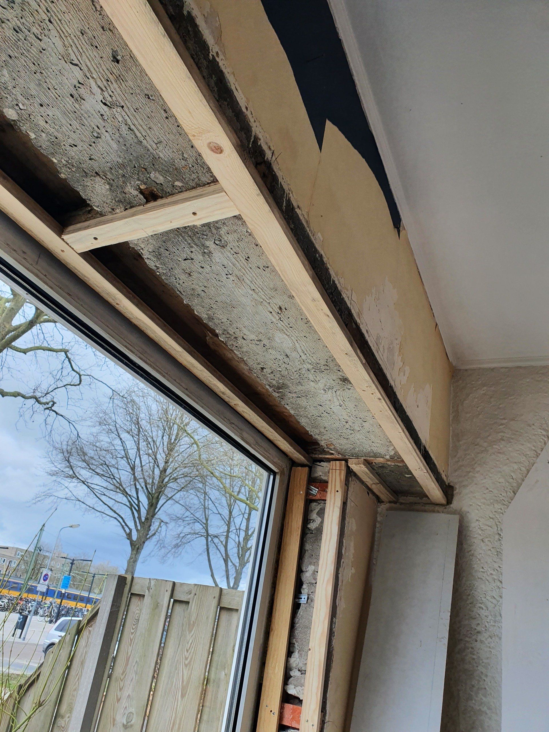 Beschadigd raamkozijn met afbladderende sierlijst en blootliggende isolatie in een kamer.