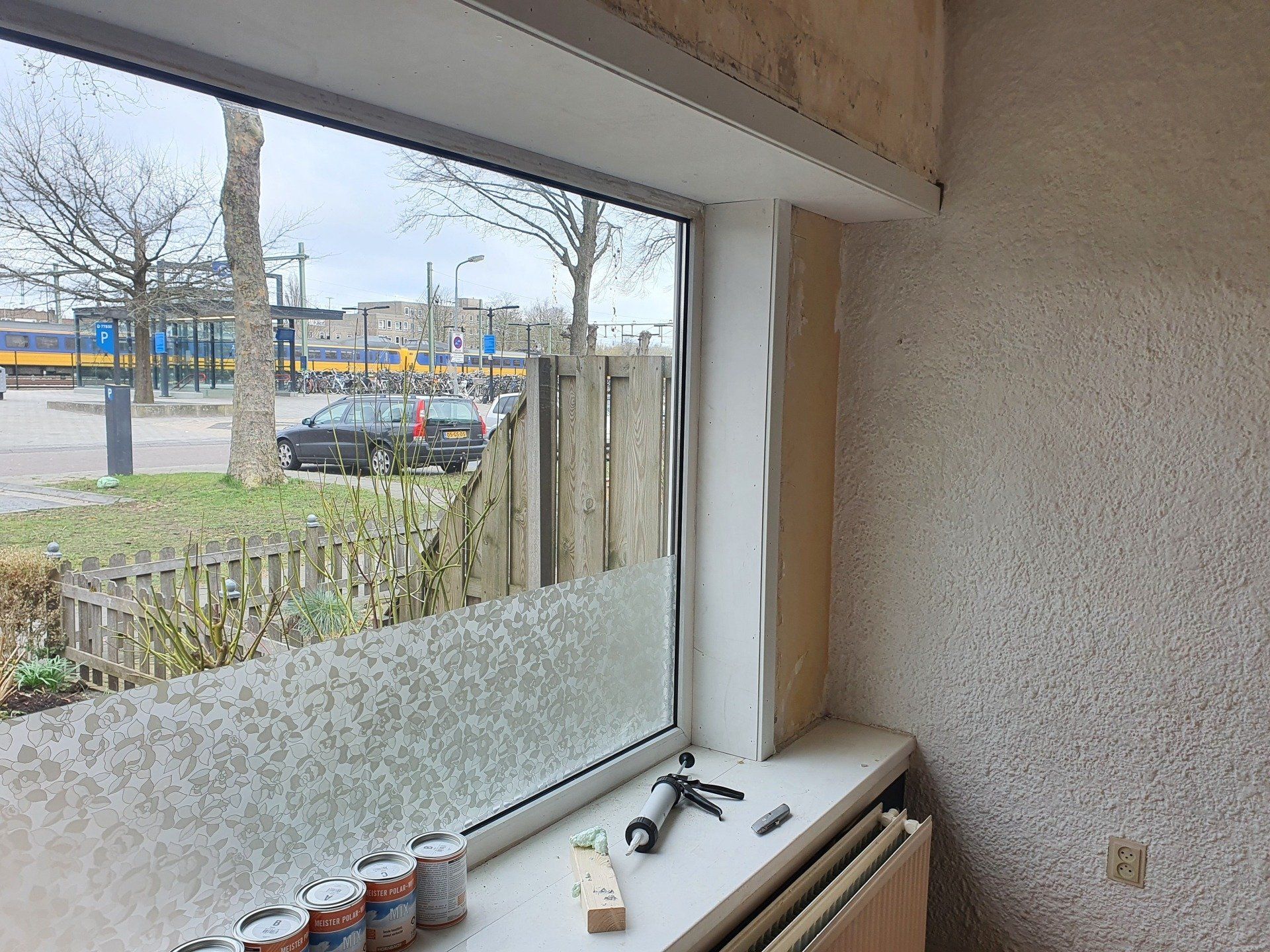 Gedeeltelijk afgewerkte kamer met matglazen raam, gereedschap op de vensterbank en uitzicht op de straat.
