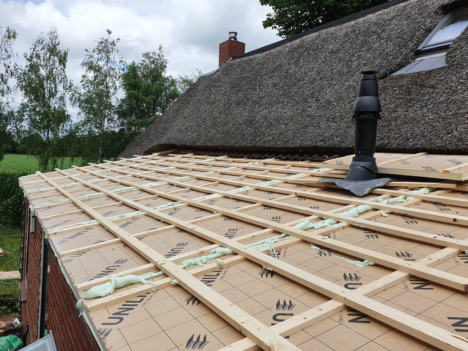 Dak in aanbouw met houten latten en een zwarte ventilatiepijp naast een huis met een rieten dak.