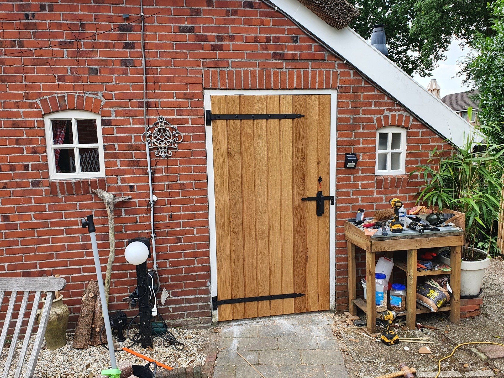 Houten tuinschuurdeur in een rode bakstenen muur, met planken en gereedschap ernaast.