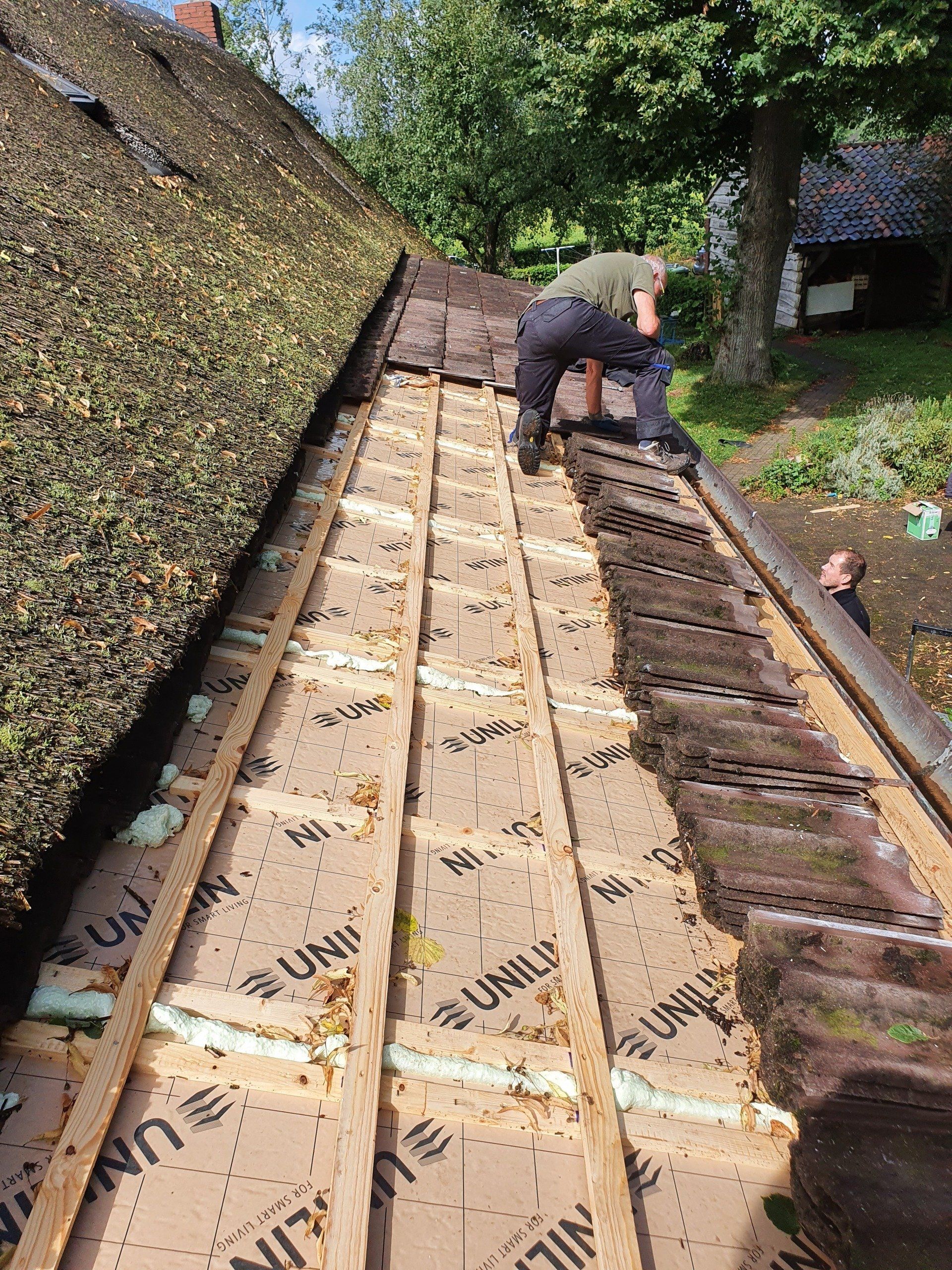 Een dakwerker is bezig met dakreparaties en knielt op de blootliggende houten planken naast een met mos begroeid leien dak.