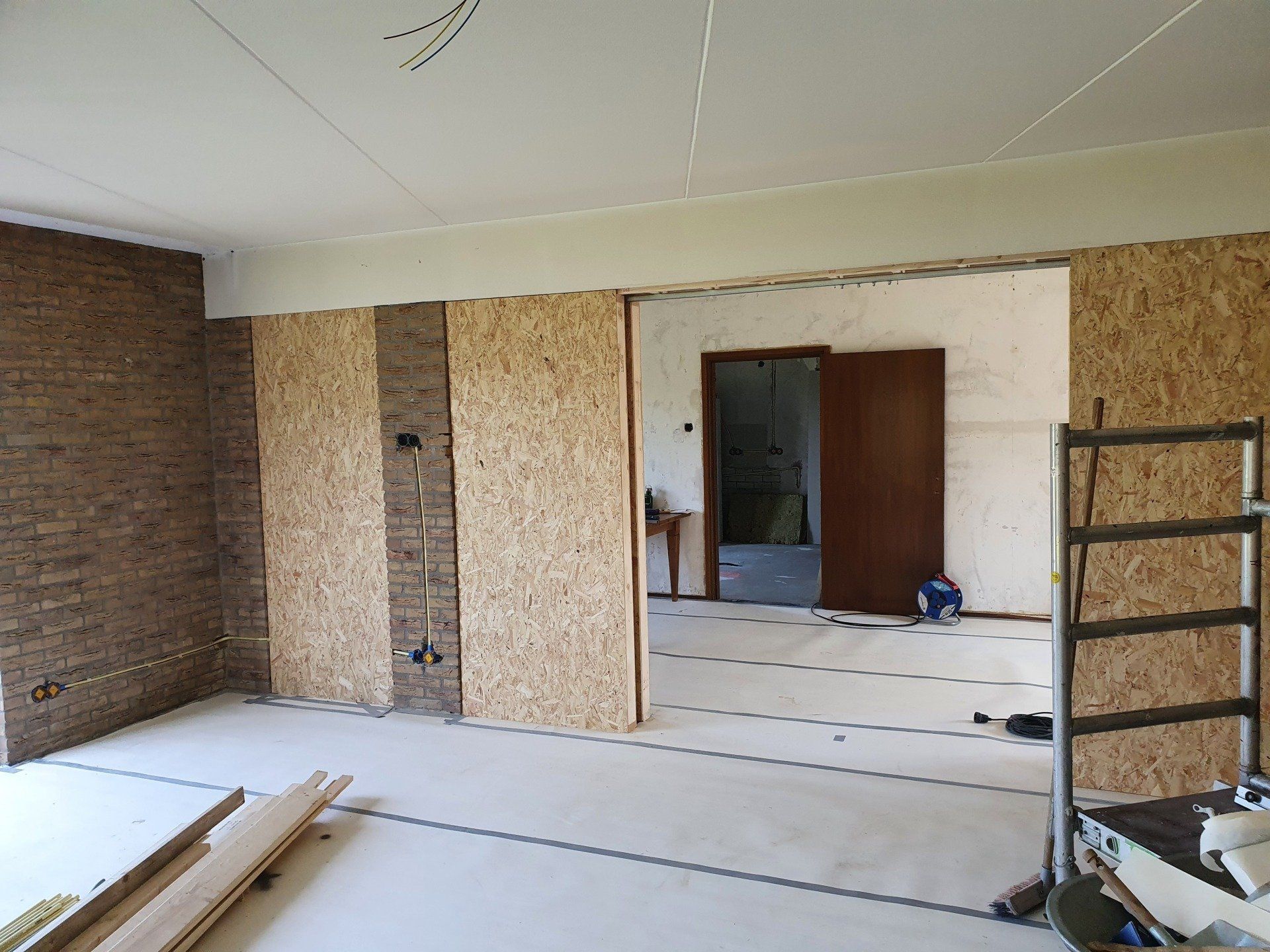 Onafgewerkte kamer met zichtbare muren, deuropening, ladder en bouwmaterialen op de vloer.