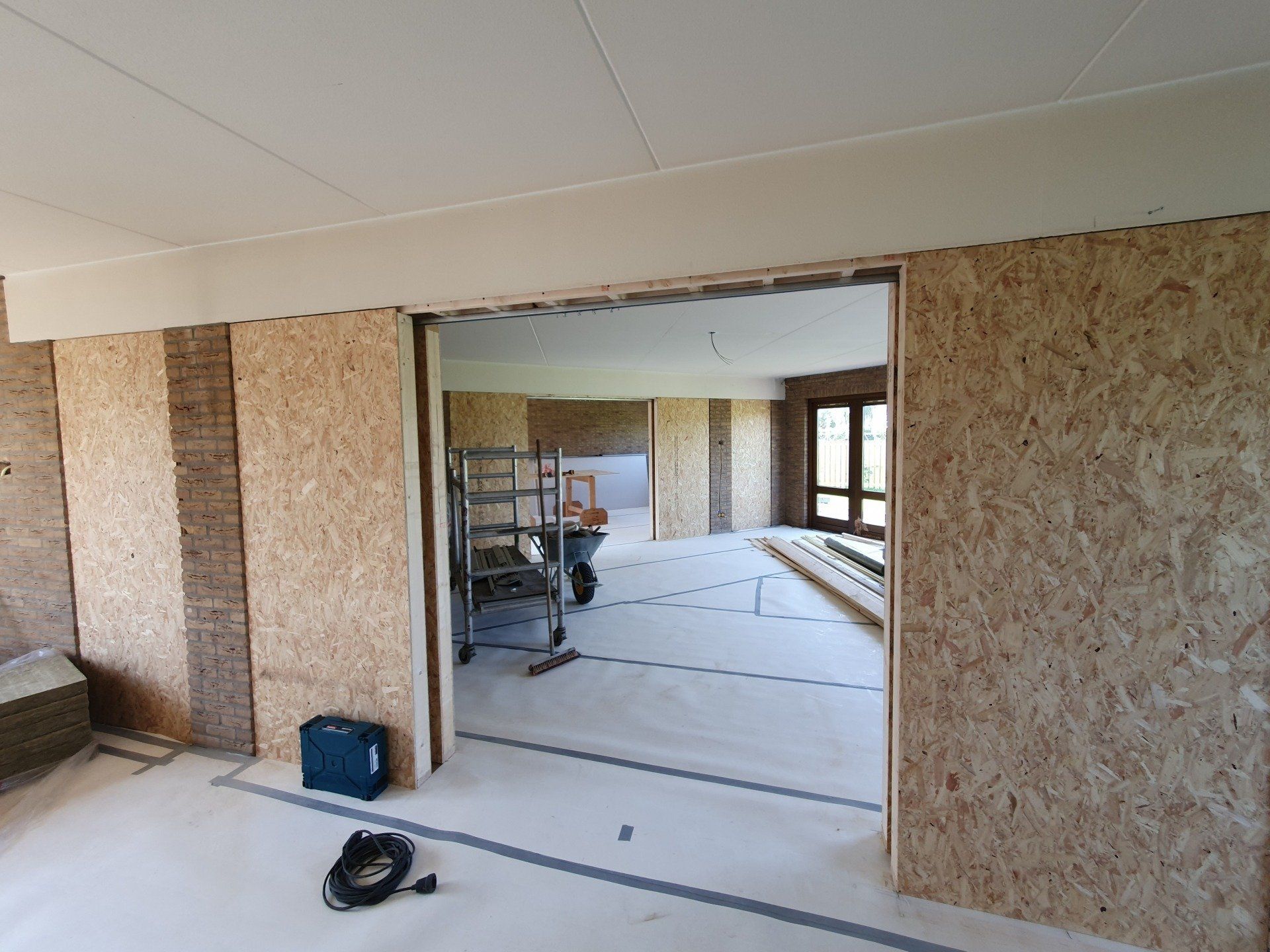 Onafgewerkte binnenruimte in renovatie, met OSB-wandpanelen, gereedschap en een open deuropening naar een andere ruimte.