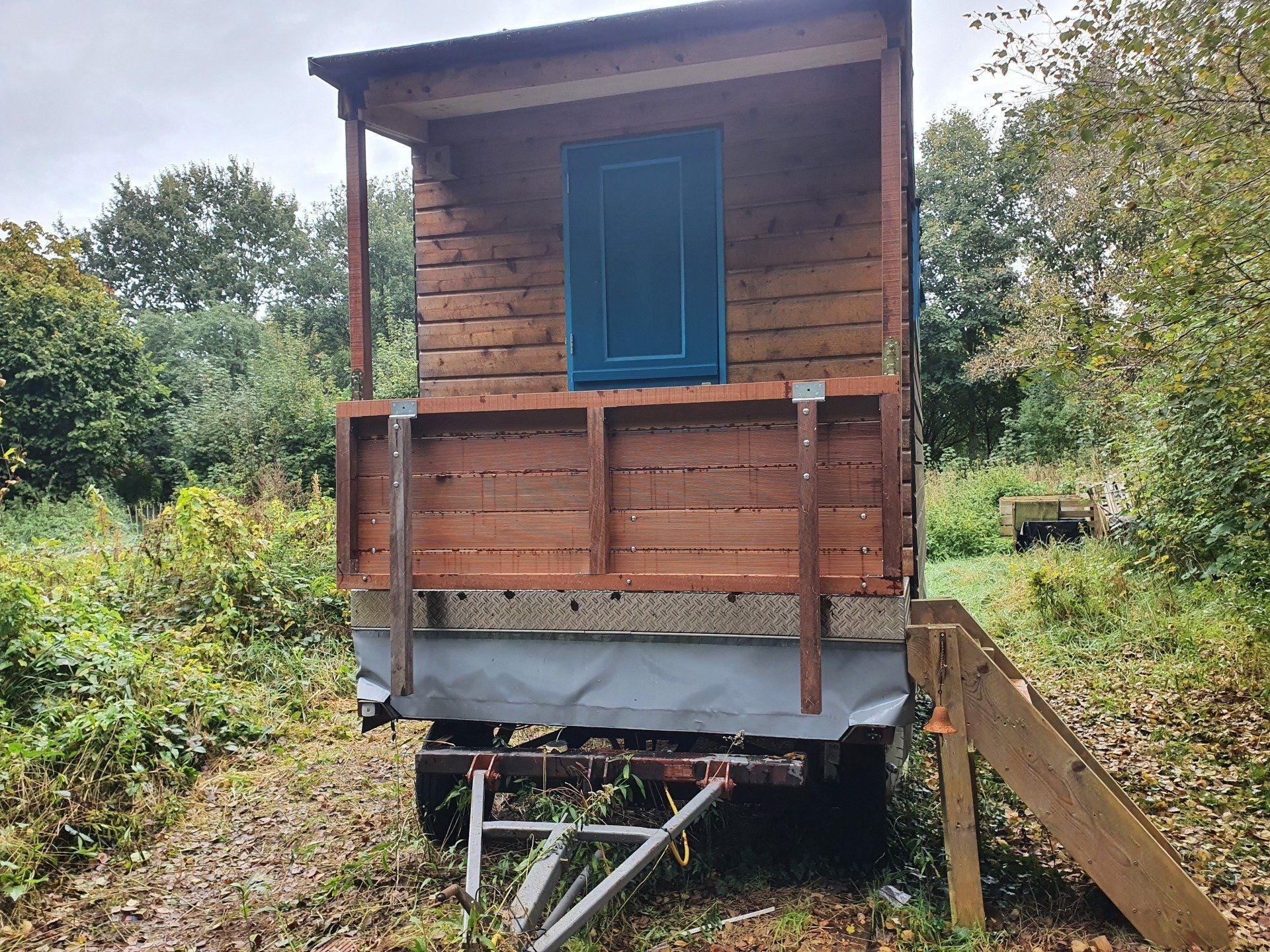 Kleine houten hut met blauwe deur op een aanhanger in een grasrijke buitenomgeving.