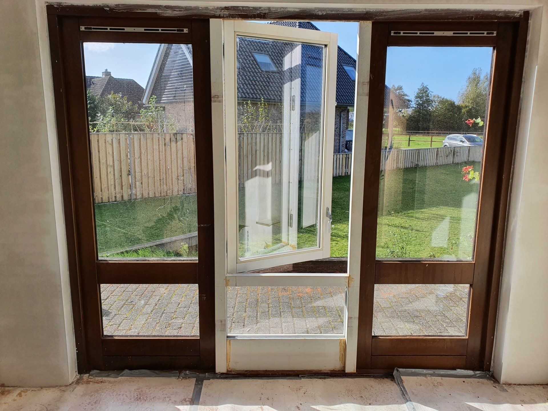 Een open witte deur in een glazen wand met houten frame biedt uitzicht op een achtertuin en een schutting.