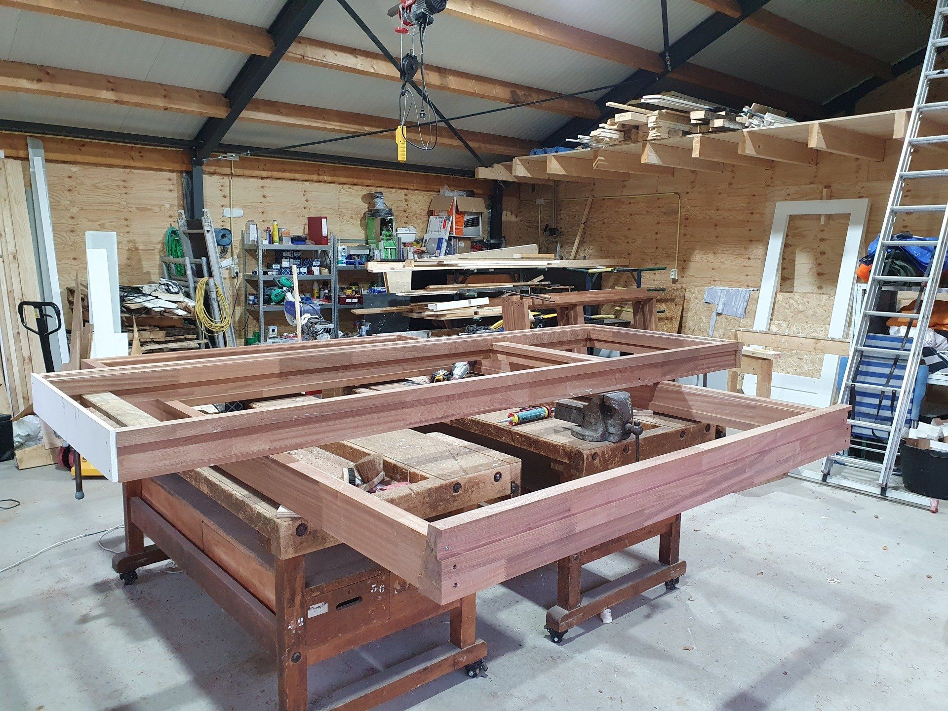 Werkplaats met een groot houten frame op schragen, omringd door gereedschap en opbergplanken.