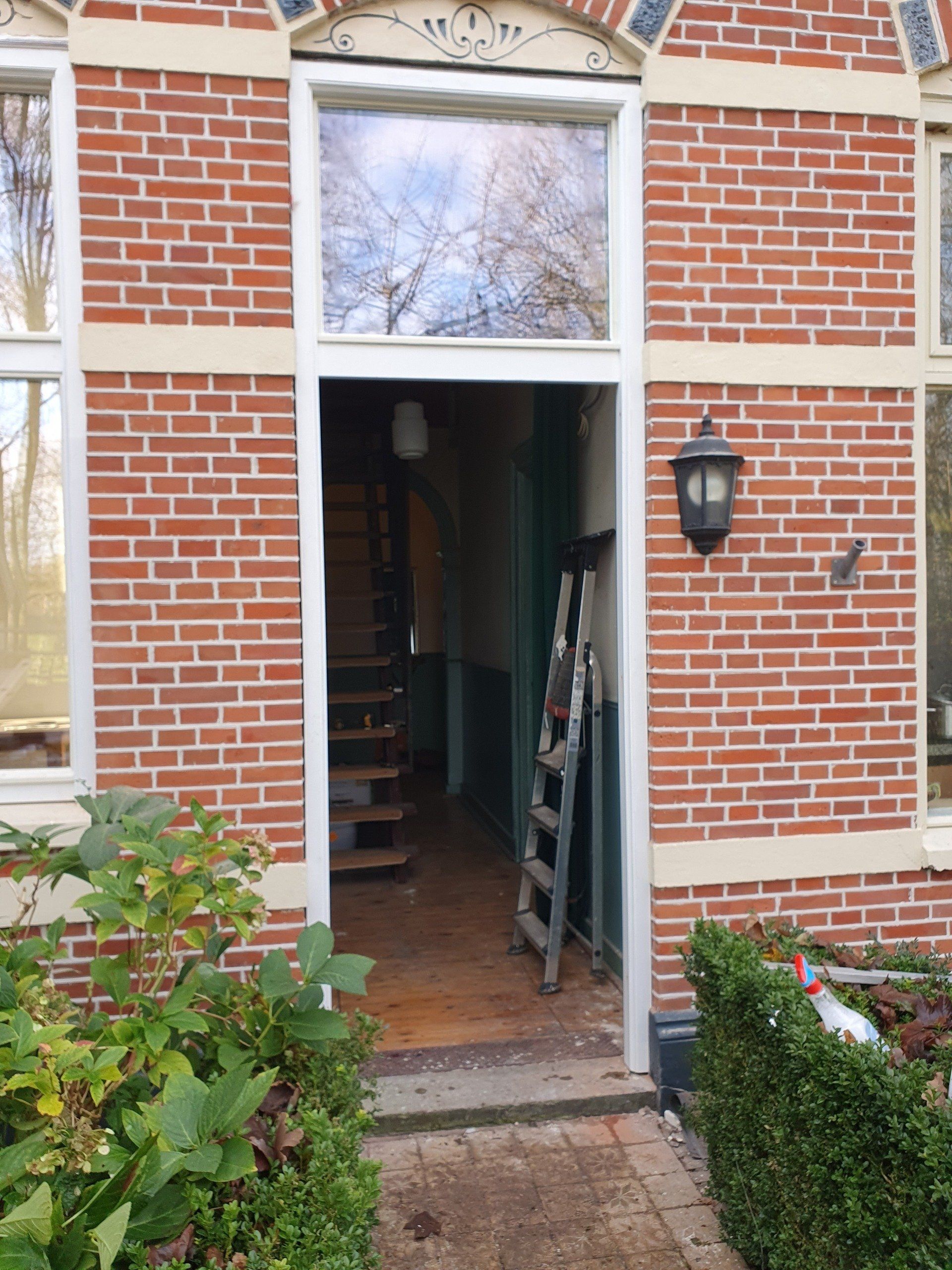 Entree van een bakstenen herenhuis met open deuropening, bakstenen pad en aan de muur gemonteerde lantaarn.