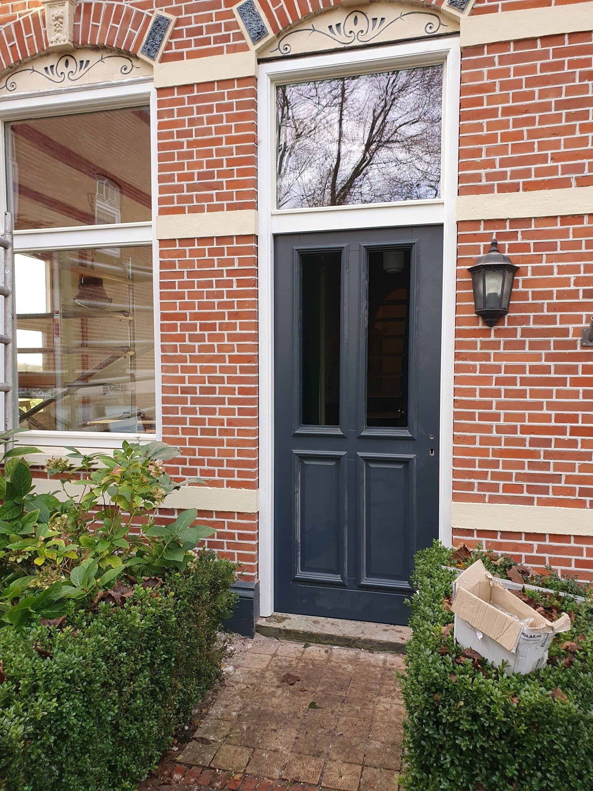 Donkerblauwe voordeur in een rood bakstenen huis met een boogvormig raam erboven en een veranda-lamp ernaast.