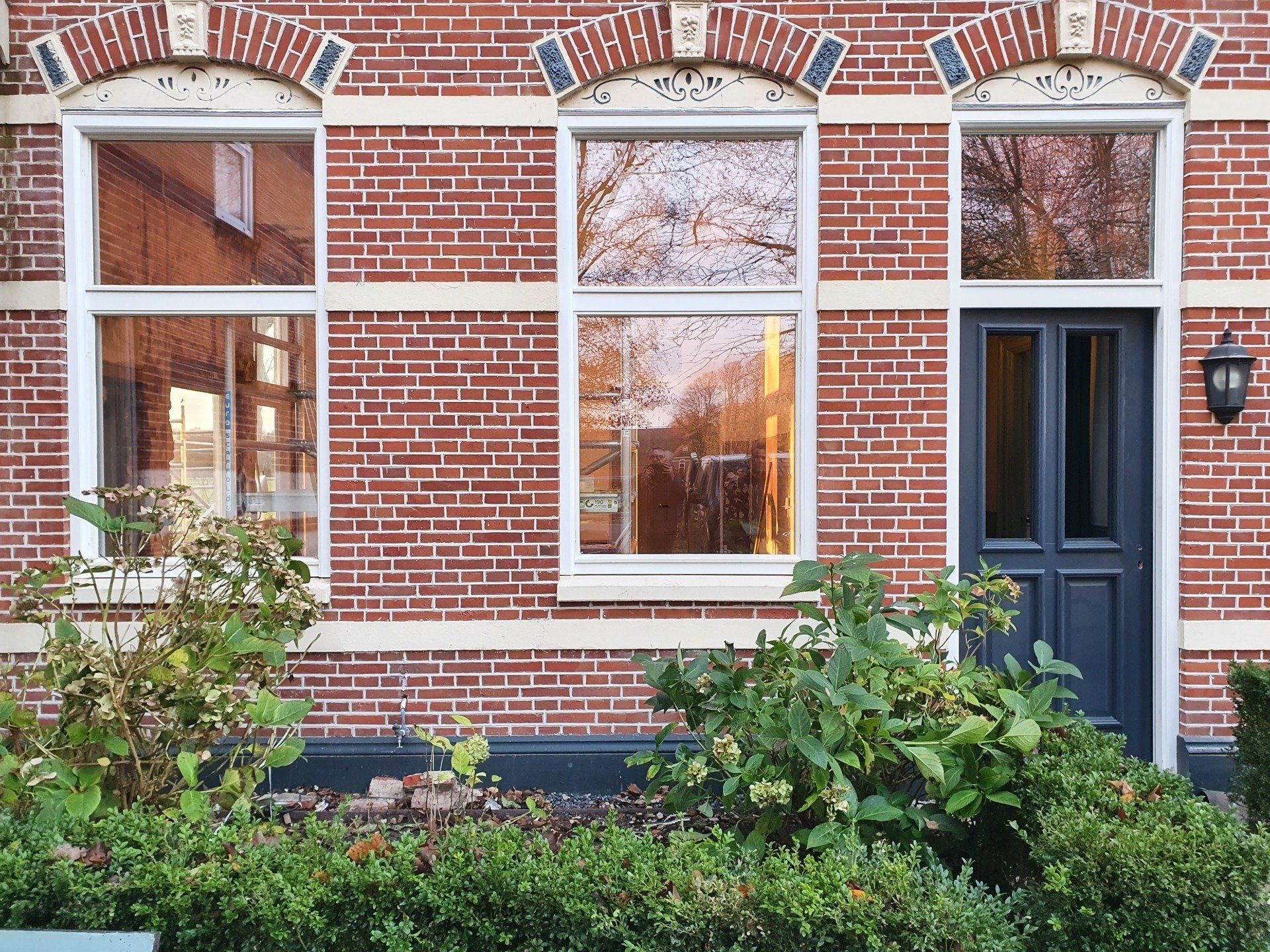 Een rijtjeshuis met een bakstenen gevel, een zwarte deur, grote ramen en groene struiken langs de voortuin.