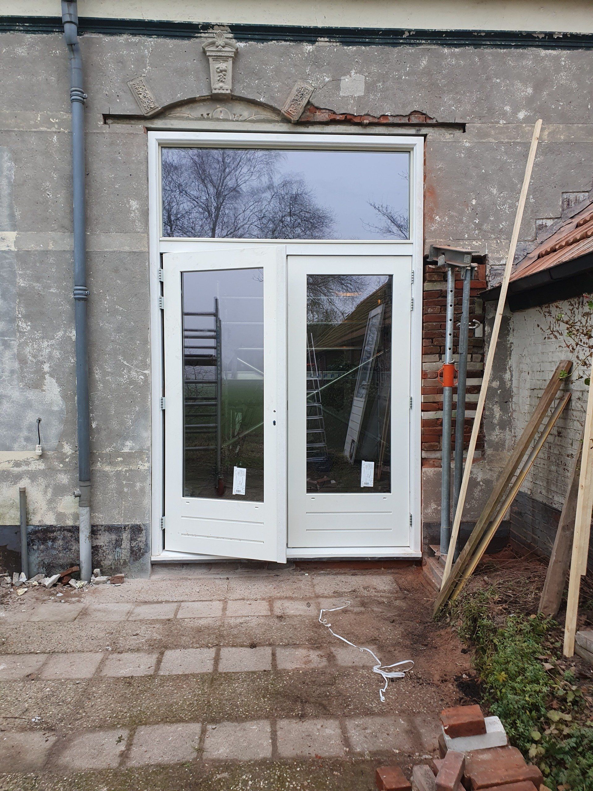 Een witte dubbele glazen deur in een ruwe buitenmuur, met een bakstenen pad en gereedschap in de buurt.