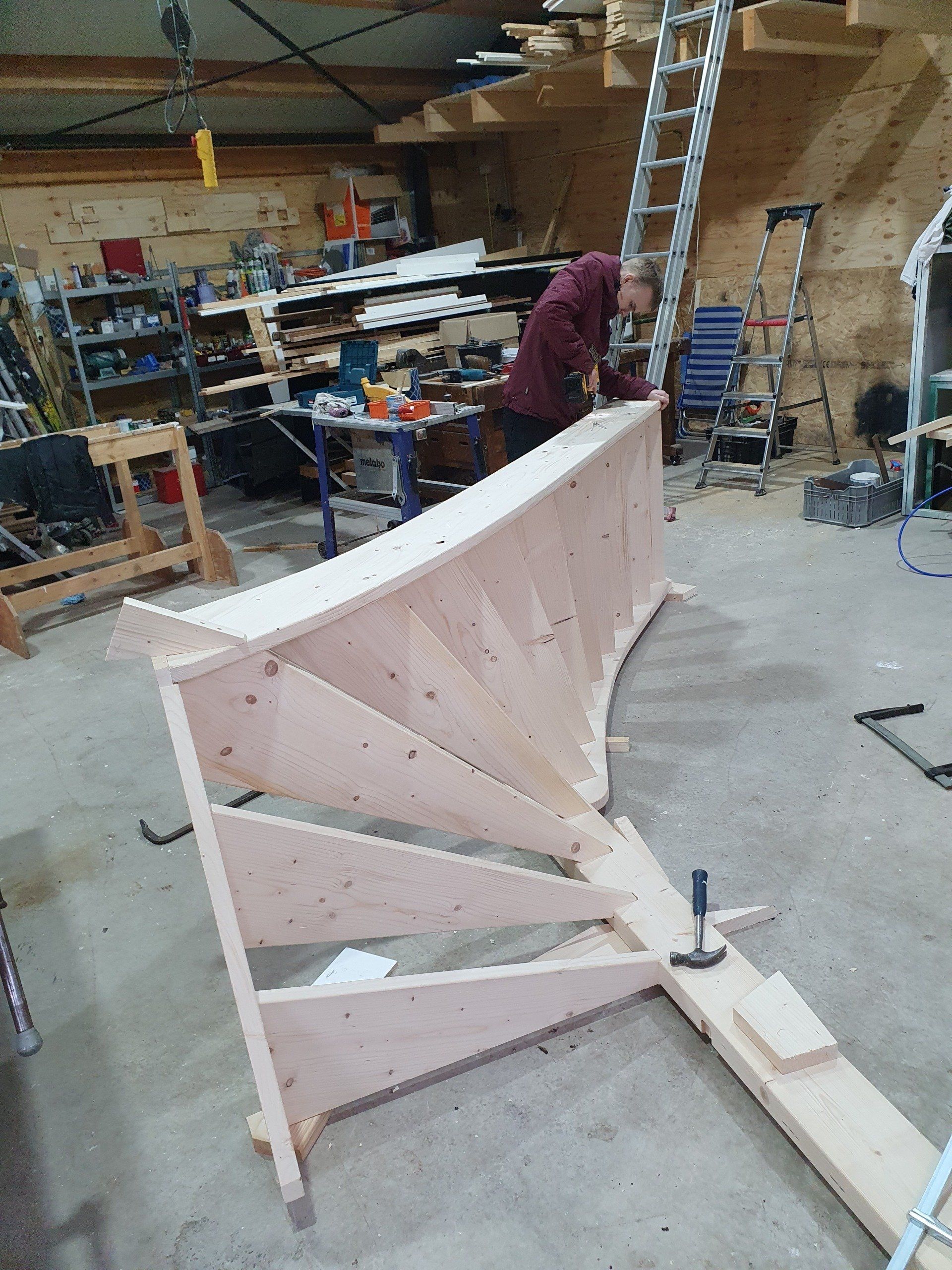 In een werkplaats wordt een houten trapconstructie gebouwd, met op de achtergrond een persoon aan het werk.