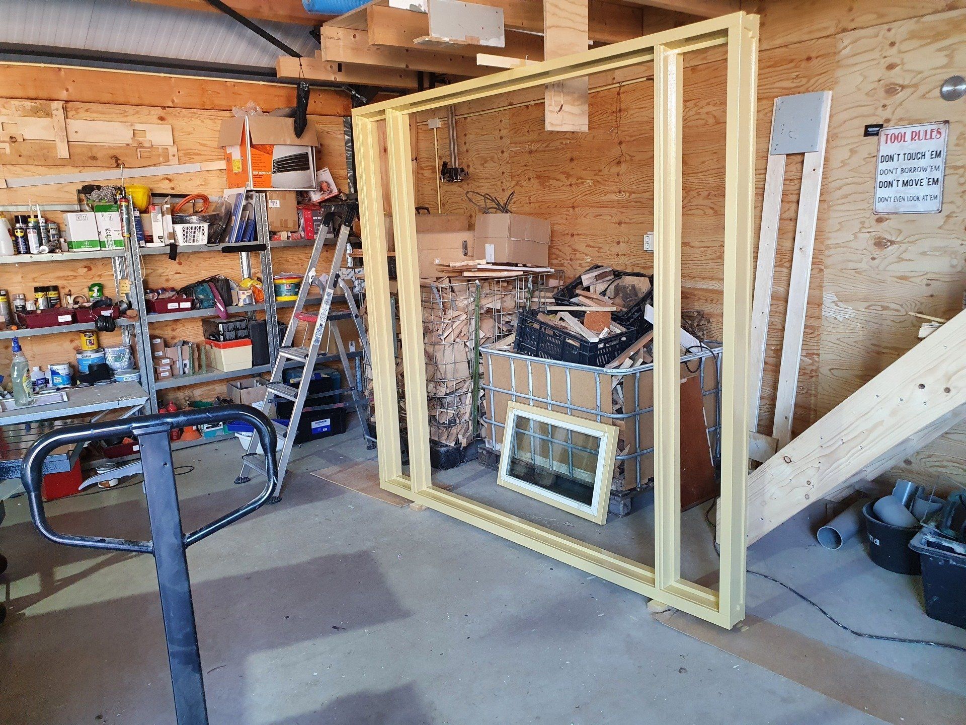 Houten frame voor een muur in een werkplaats/garage, met gereedschap en planken op de achtergrond.