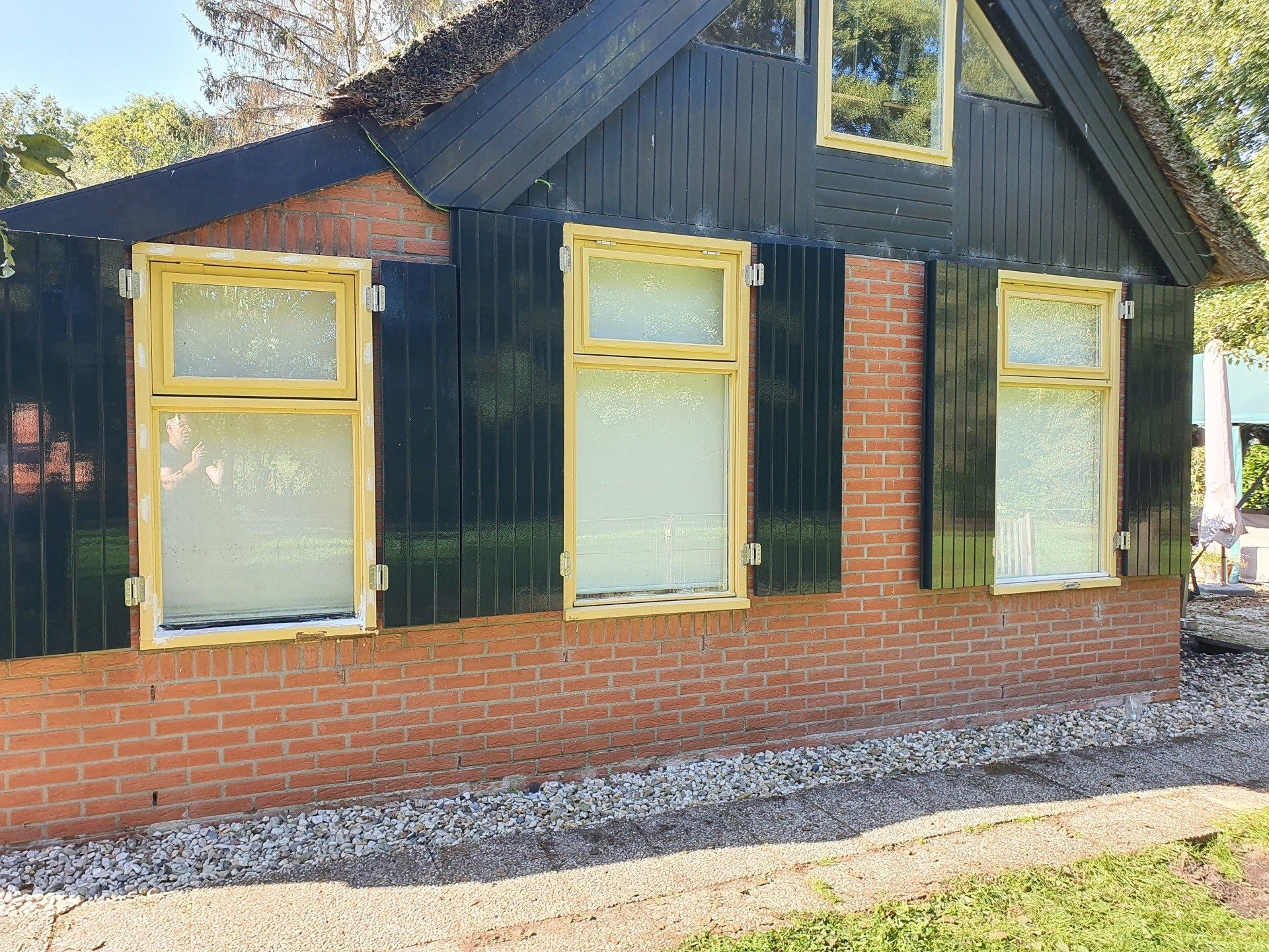 Klein bakstenen huisje met zwarte gevelbekleding, geel omlijste ramen en een zadeldak, badend in het daglicht.