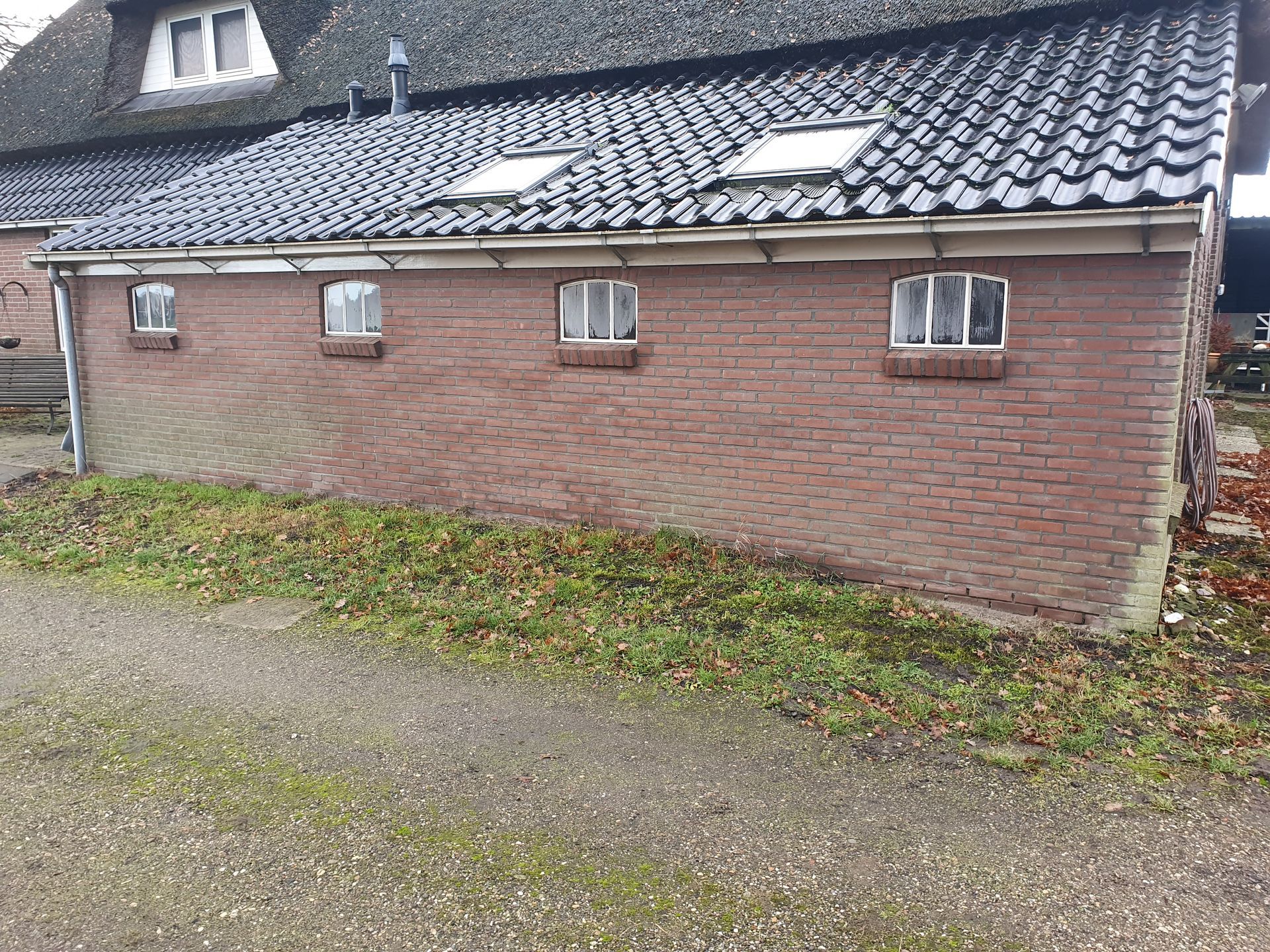 Een stenen bijgebouw met een donker pannendak en kleine ramen, gelegen naast een grindpad.