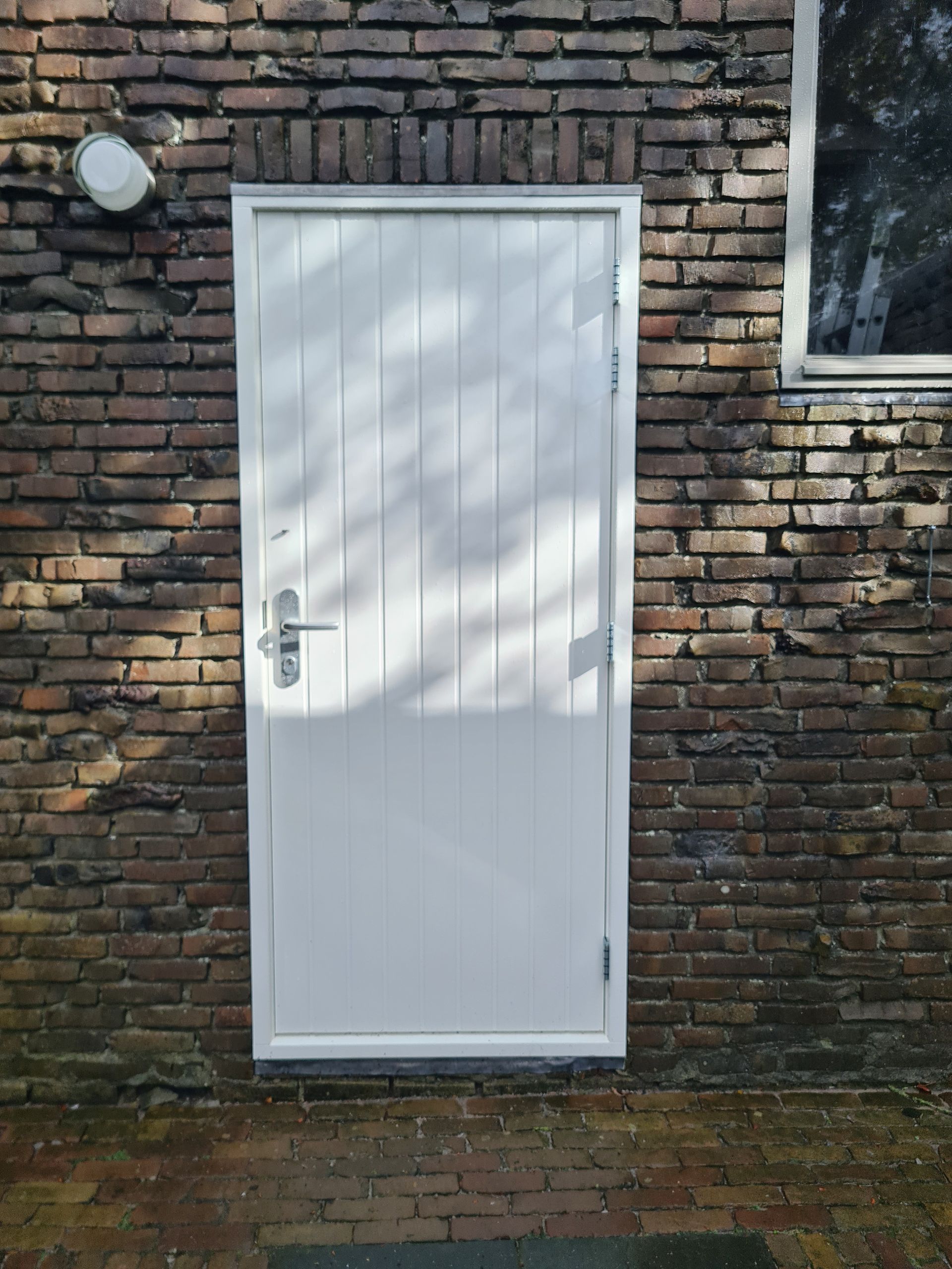 Witte deur met metalen handgreep, ingebouwd in een donkere bakstenen buitenmuur.